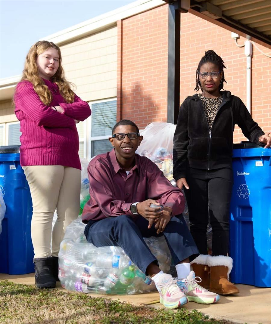 Three teenagers pose with recycling bins and a bag of plastic bottles outside a brick building.